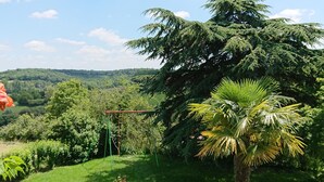 A lush garden with a swing set, a palm tree, and a view of a forested hill. - Cottage Le Breuil in Périgord - Charm and Comfort (Salles-Lavalette)
