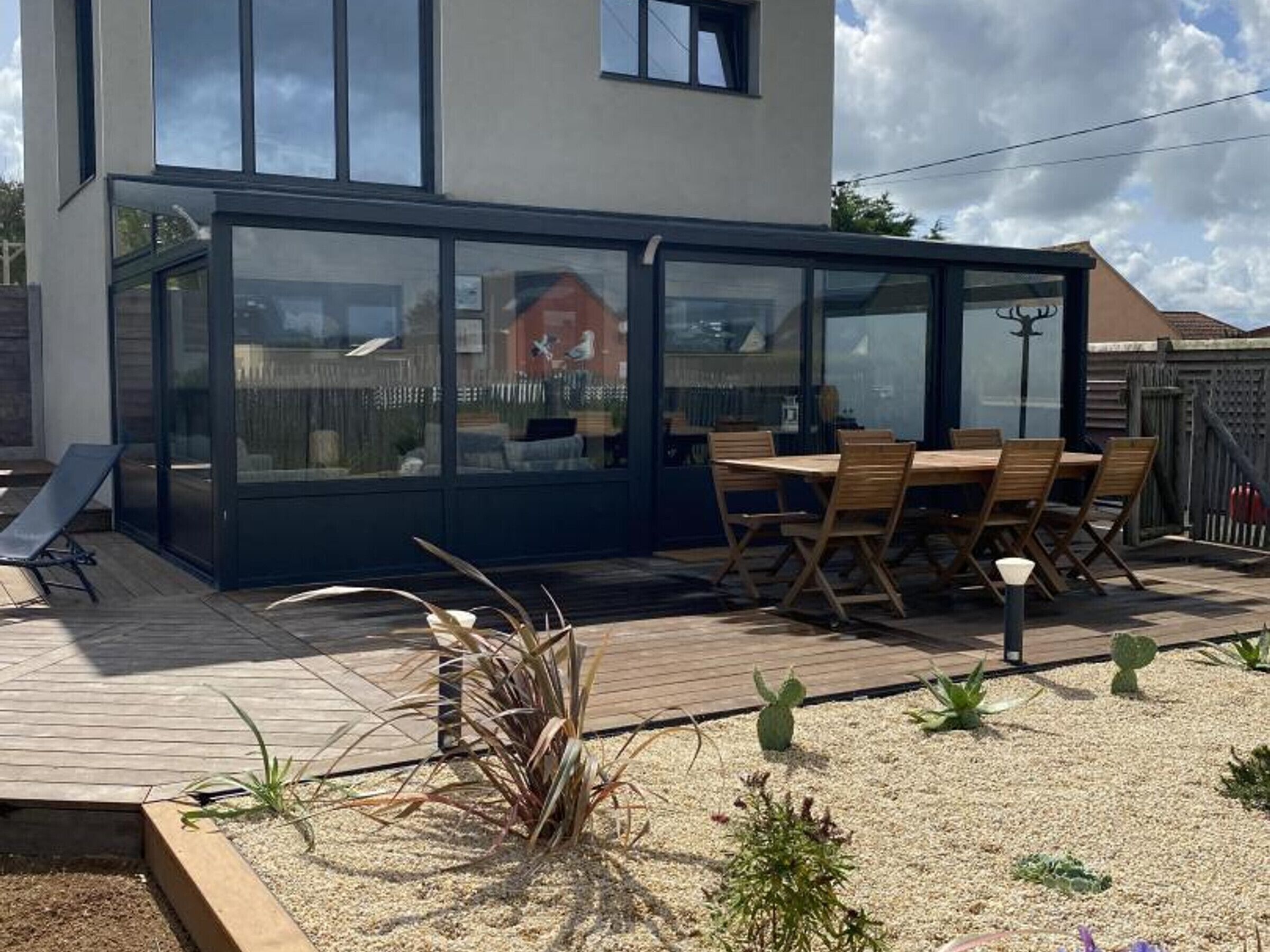 A modern house with a glass-enclosed patio area, a wooden deck, and a dining table with chairs.