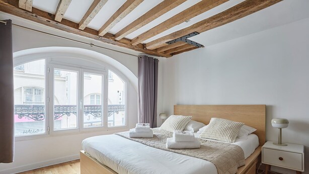 A bedroom with a wooden bed, a nightstand, and a window with a view of buildings. - Charming Apartment Center of Paris (Paris)