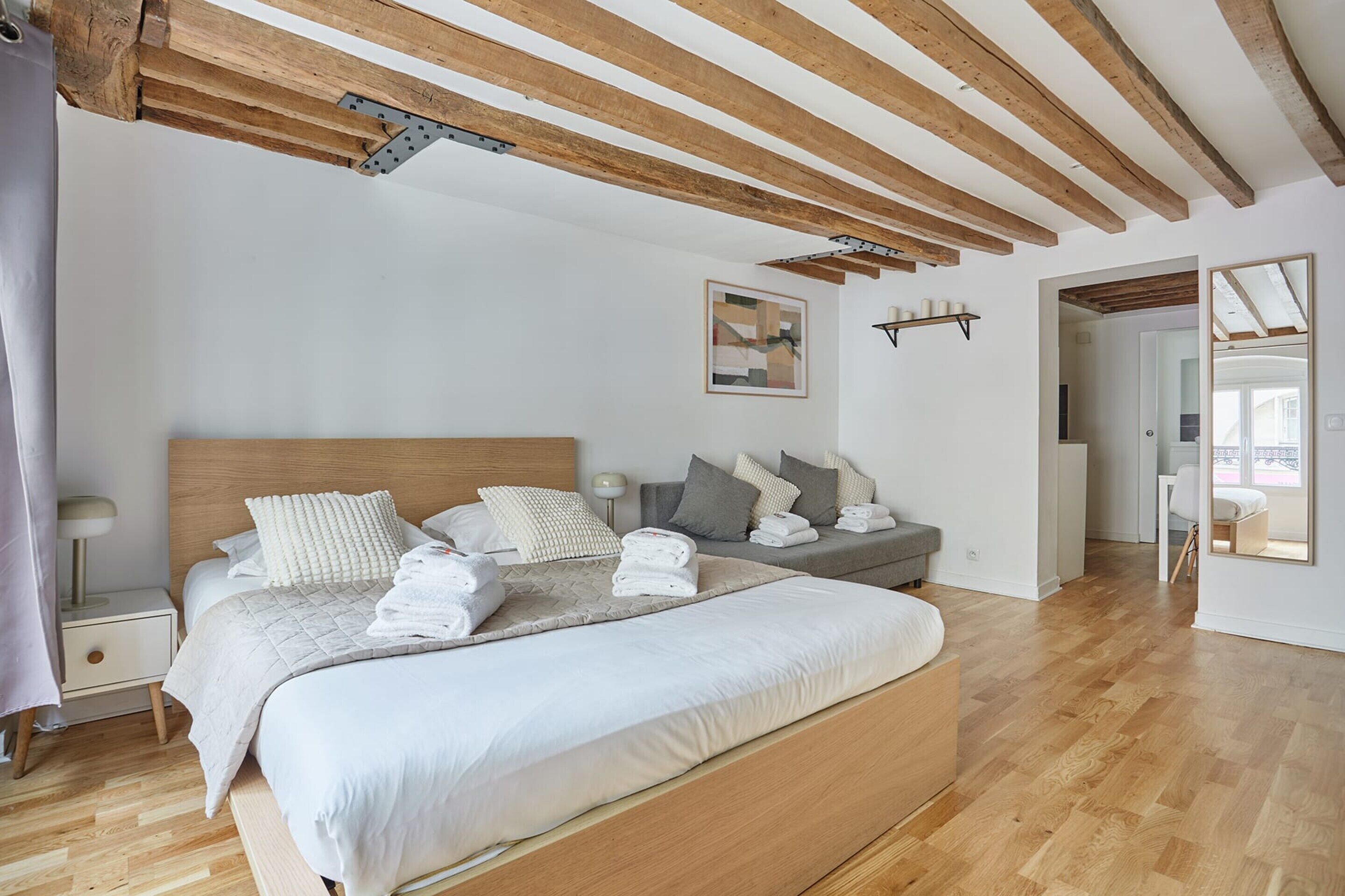 A bedroom with a large bed, wooden beams, a nightstand, a bedside table, a wall-mounted shelf, and a door leading to another room.