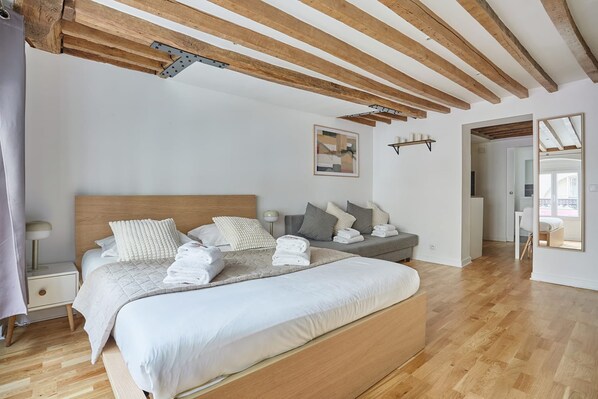 Een slaapkamer met een groot bed, houten balken, een nachtkastje, een wandplank en een deur naar een andere kamer.