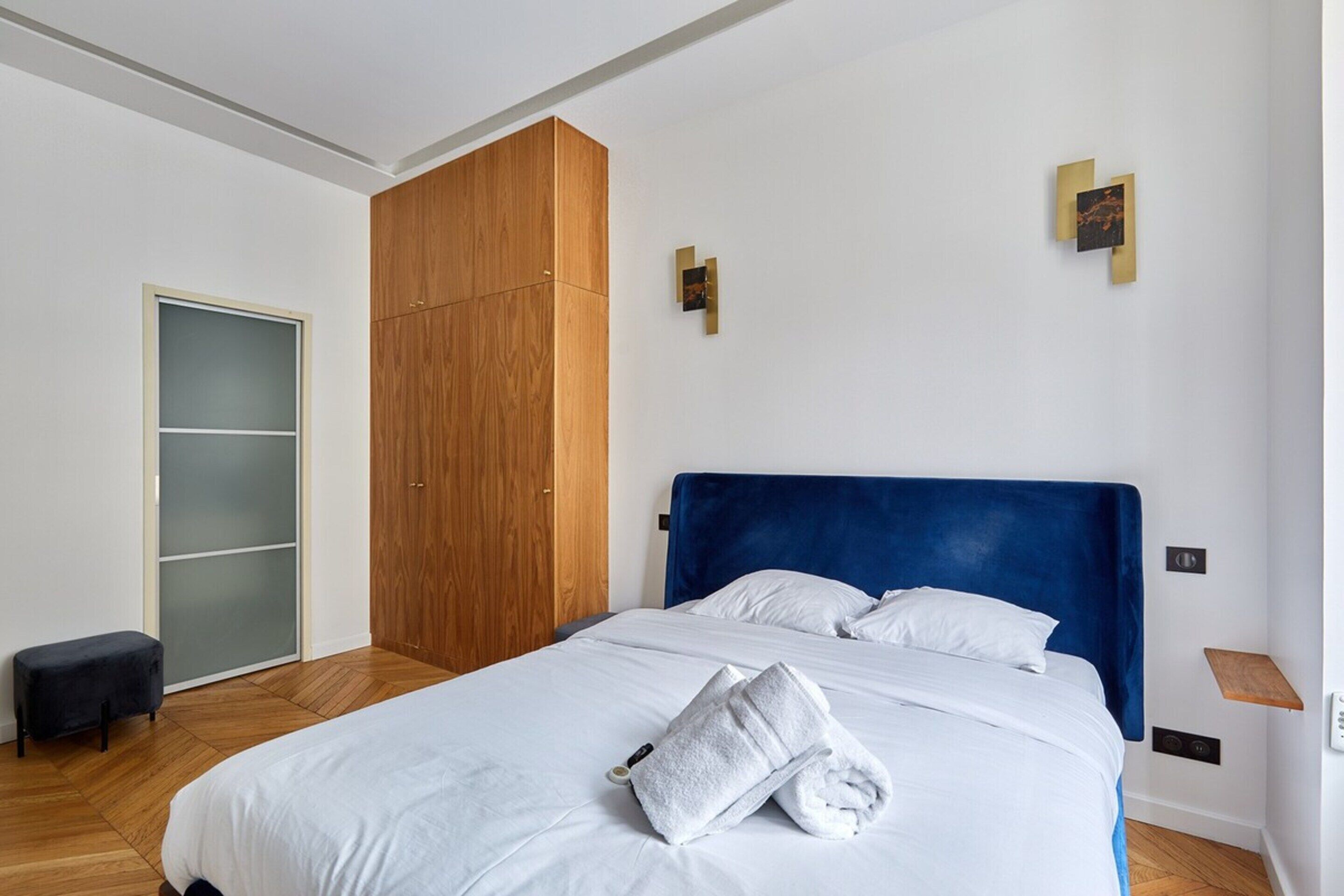 A modern bedroom with a large bed, wooden wardrobe, and a small stool.