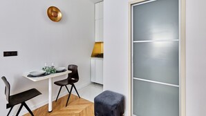 A modern dining area with a white table, two black chairs, a small round table with a plant, and a grey cushioned stool. - Cosi Apartment Bastille-le Marais (Paris)