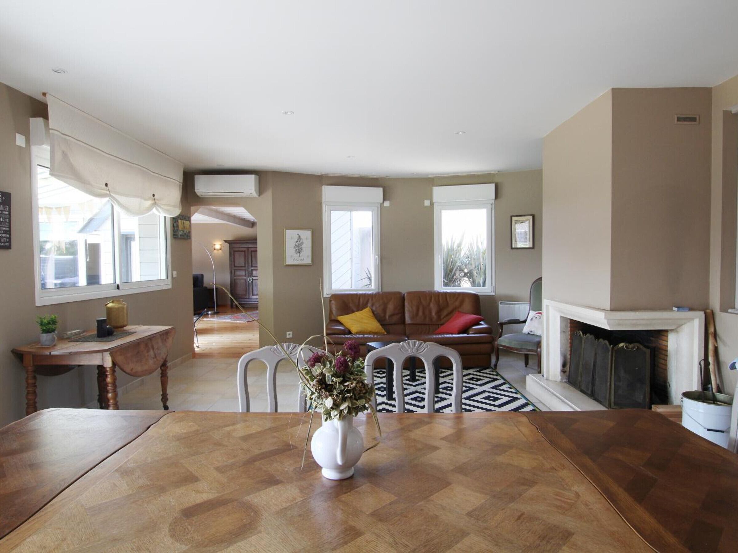 Un vaste salon comprenant une table à manger en bois, une cheminée et un coin salon confortable avec un canapé en cuir marron et un tapis à motifs.