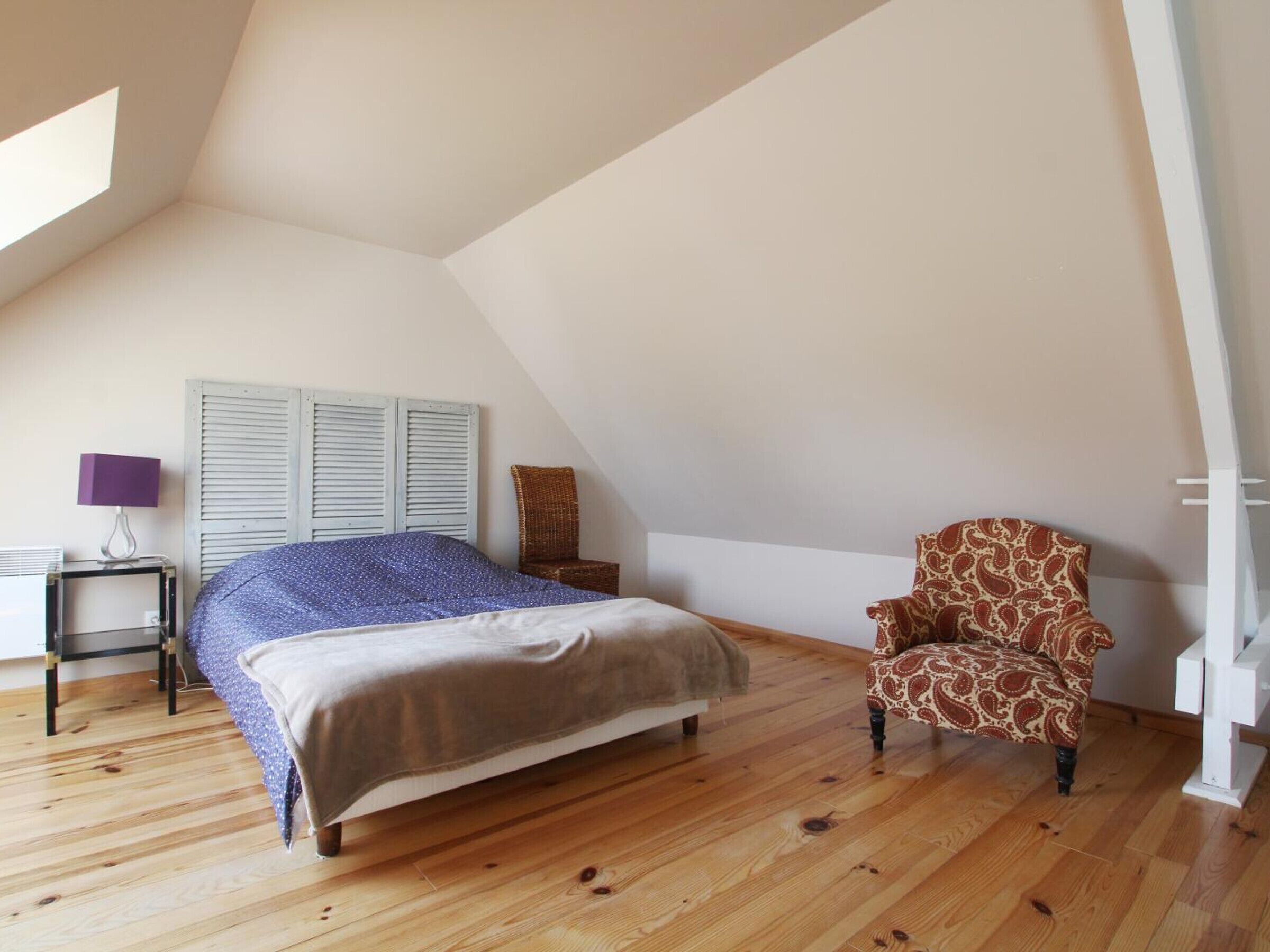 Une chambre avec un plafond en pente, un lit recouvert d’une couvre-lit à motifs, une chaise ornée d’un motif cachemire, une petite table avec une lampe et une armoire blanche.