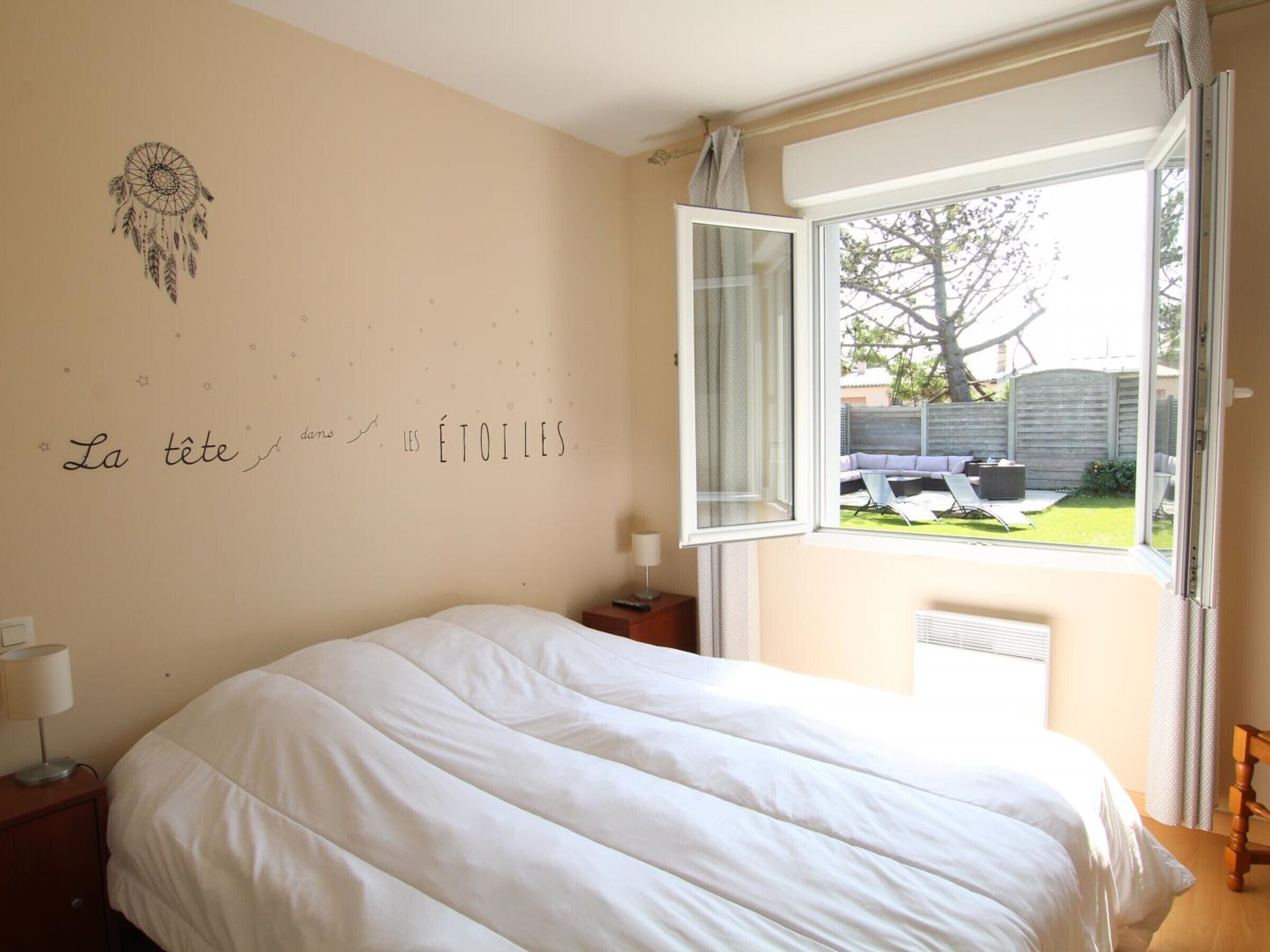 A bedroom with a bed, a nightstand, a lamp, a window with a view of the outside, and a wall with a dreamcatcher and text.