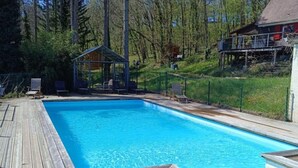 Una piscina rettangolare con un solarium in legno, circondata da una recinzione e alberi.
