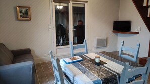 Un'area pranzo con un tavolo apparecchiato per due, un divano, una televisione a parete e una finestra con tende.