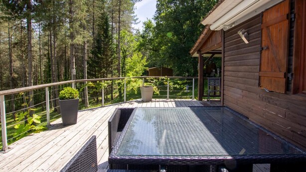 Un patio in legno con tavolo e sedie in vetro, immerso nel verde degli alberi.