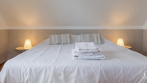 Een net opgemaakt bed met witte lakens en een opgevouwen handdoek, twee bedlampen, en een gestructureerde muur.