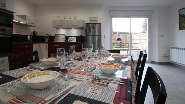 Une cuisine moderne équipée d’appareils électroménagers en acier inoxydable, un coin repas avec une table dressée pour six personnes, et une vue sur le jardin à travers une porte-fenêtre coulissante.