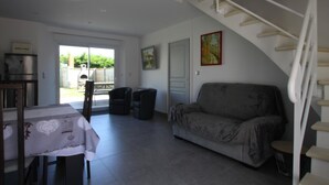 Un salon avec un canapé gris, une table à manger recouverte d’une nappe, un escalier et une porte donnant sur l’extérieur.