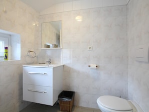 A bathroom with white marble tiles, a white sink with a mirror above it, a toilet, and a trash bin. - Cozy house in Auderville with sea view, near trails (La Hague)