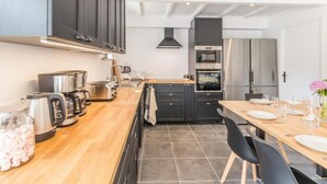 A modern kitchen with dark cabinets, stainless steel appliances, and a wooden dining table set for six. - Spacious house with heated pool and garden, near beaches (Bricqueville-sur-Mer)