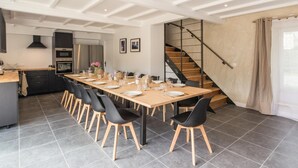 A modern kitchen with dark cabinets and stainless steel appliances, a dining area with a long table set for six, and a staircase leading to an upper level. - Spacious house with heated pool and garden, near beaches (Bricqueville-sur-Mer)