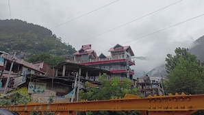 A multi-story hotel with a red and white facade, located in a mountainous area with visible greenery and clouds. - Hotel Aaditya (Kullu)