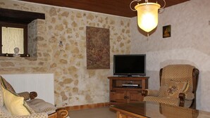 A living room with stone walls, a television on a wooden stand, two patterned armchairs, and a glass-top coffee table.