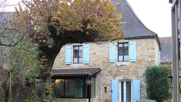 A stone house with blue shutters, a gravel driveway, and a large tree in front.