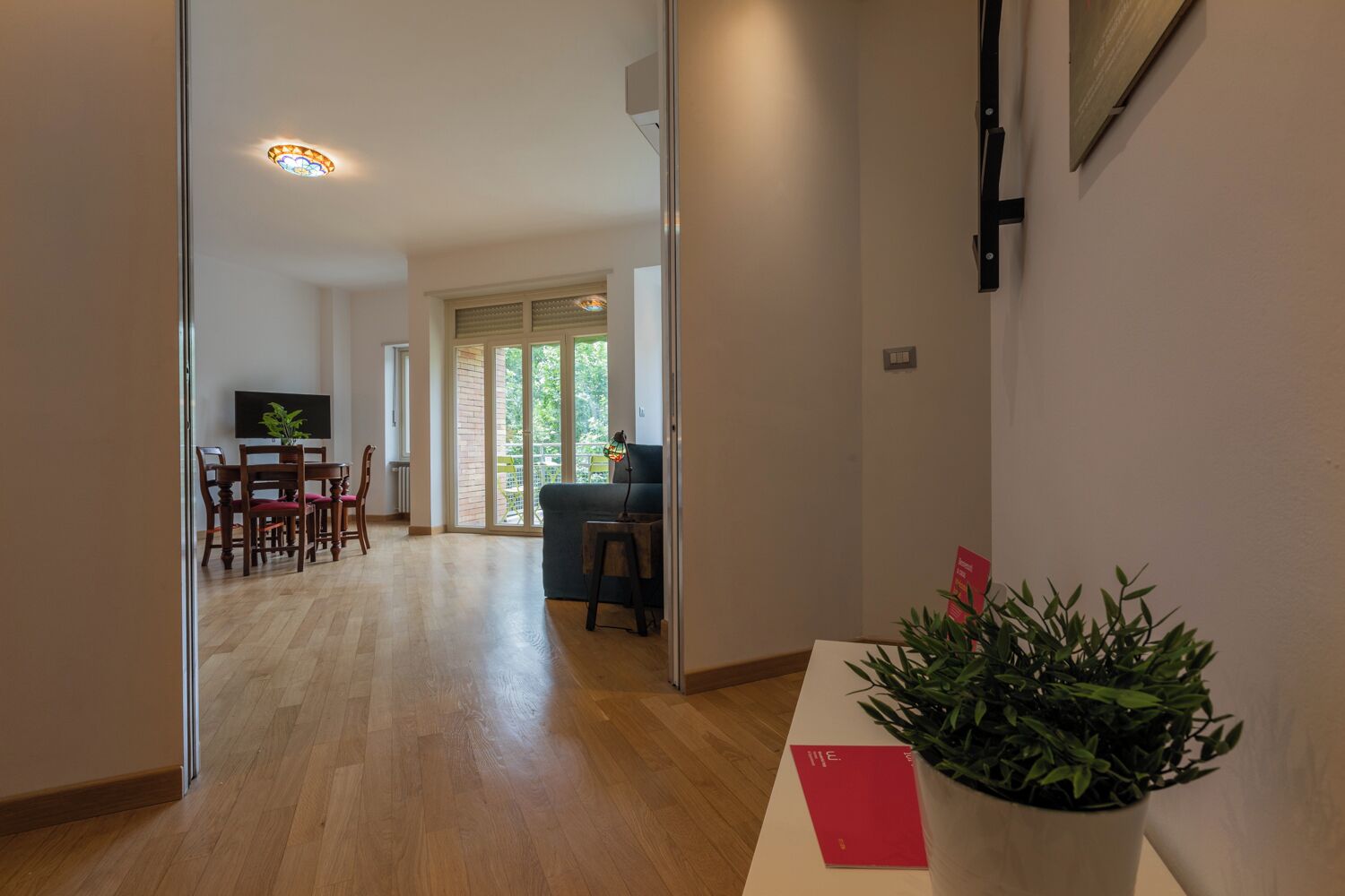 Un corridoio con pavimento in legno che conduce a una zona pranzo con tavolo e sedie, un divano e una televisione. Una pianta in vaso su un tavolo e una porta sulla destra.