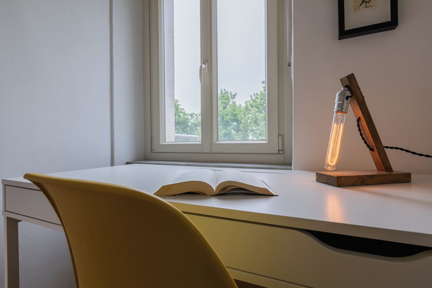 A desk with an open book, a lamp, and a window with a view of trees.