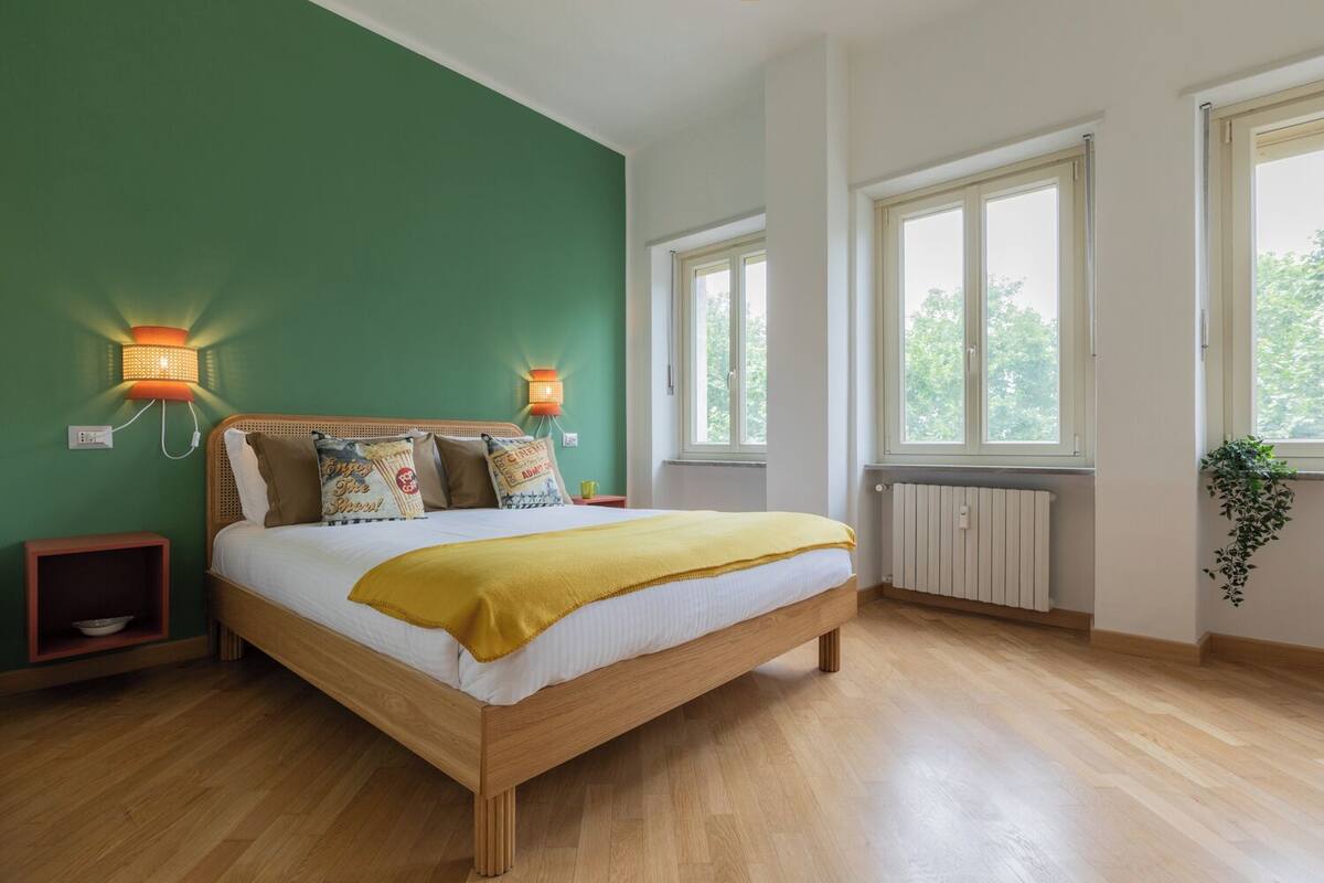 A bedroom with a wooden bed, green walls, two wall-mounted lamps, a window with a view of trees, and a radiator.