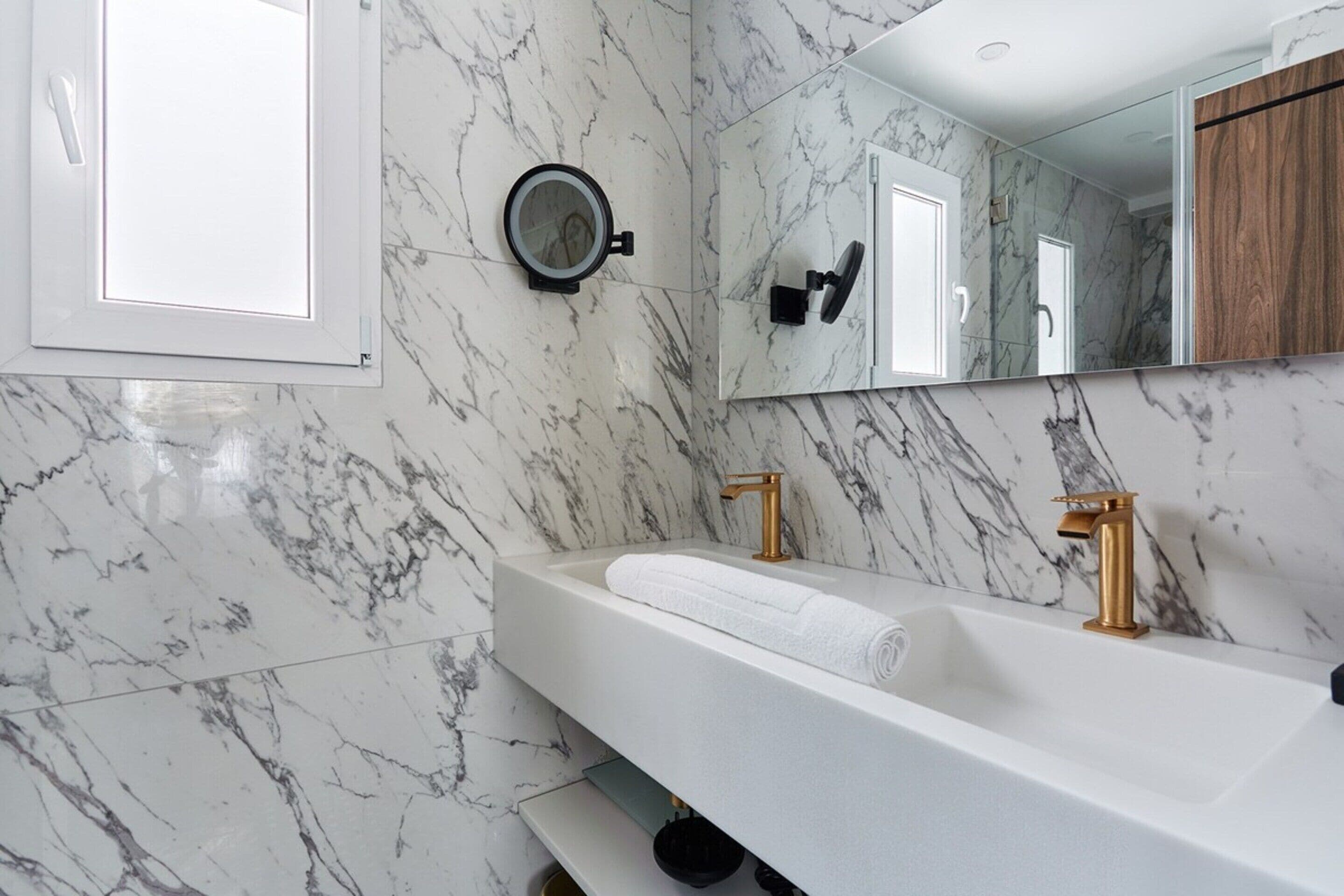 A modern bathroom with a marble backsplash, a large mirror, a white sink with gold faucets, and a towel on the counter.