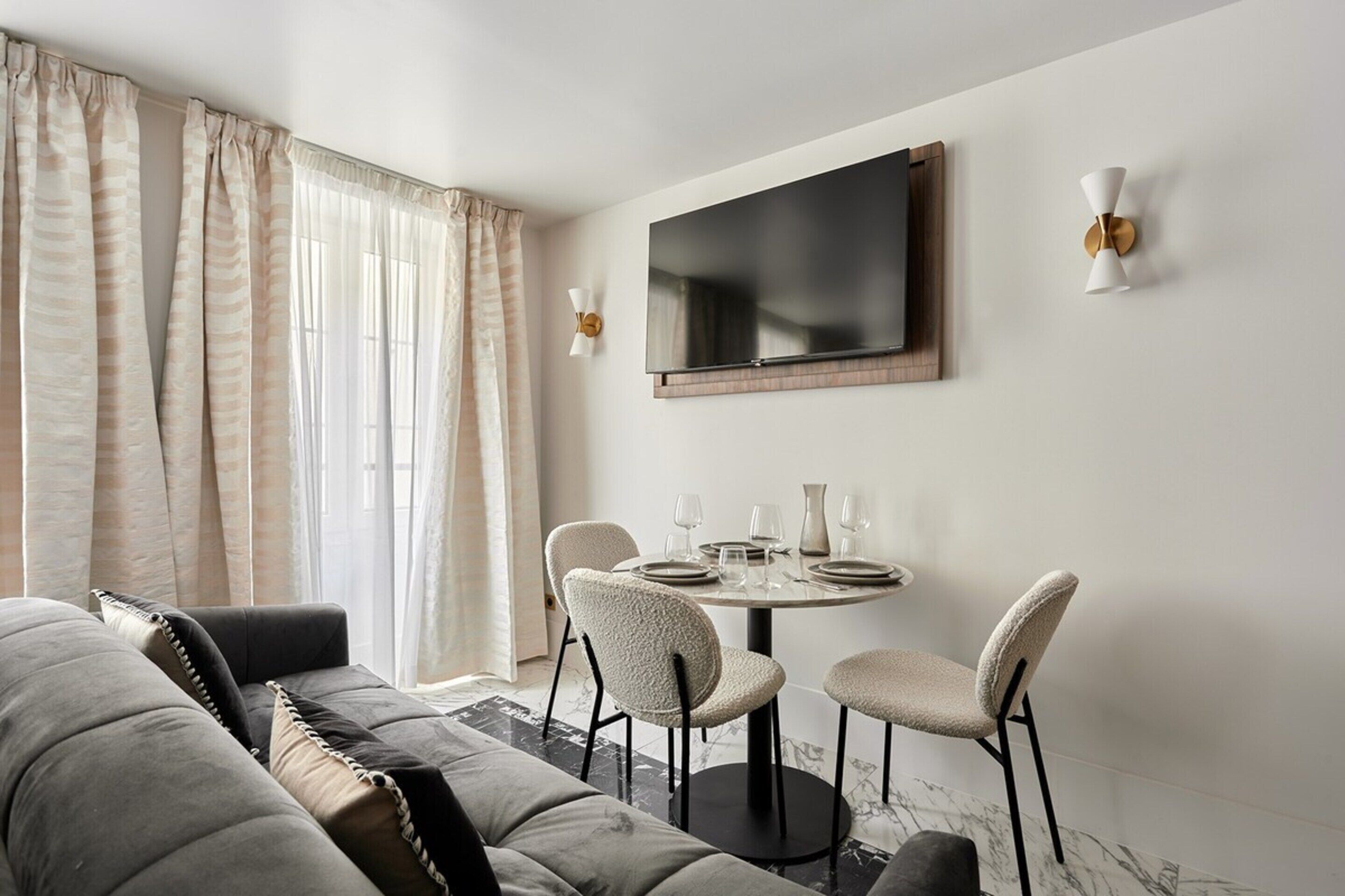 Un salon moderne comprenant un canapé, une table à manger avec des chaises, un téléviseur à écran plat fixé au mur et une grande fenêtre avec des rideaux.