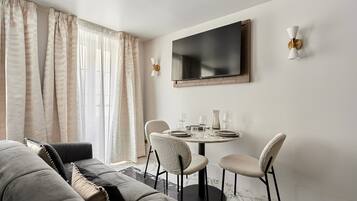 Un salon moderne comprenant un canapé, une table à manger avec des chaises, un téléviseur à écran plat fixé au mur et une grande fenêtre avec des rideaux.