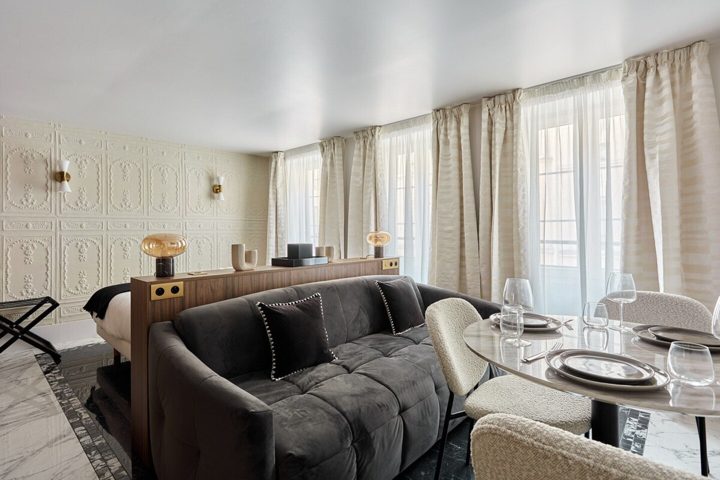 A modern living room with a dark sofa, a wooden sideboard, a round dining table with chairs, and a large window with curtains.