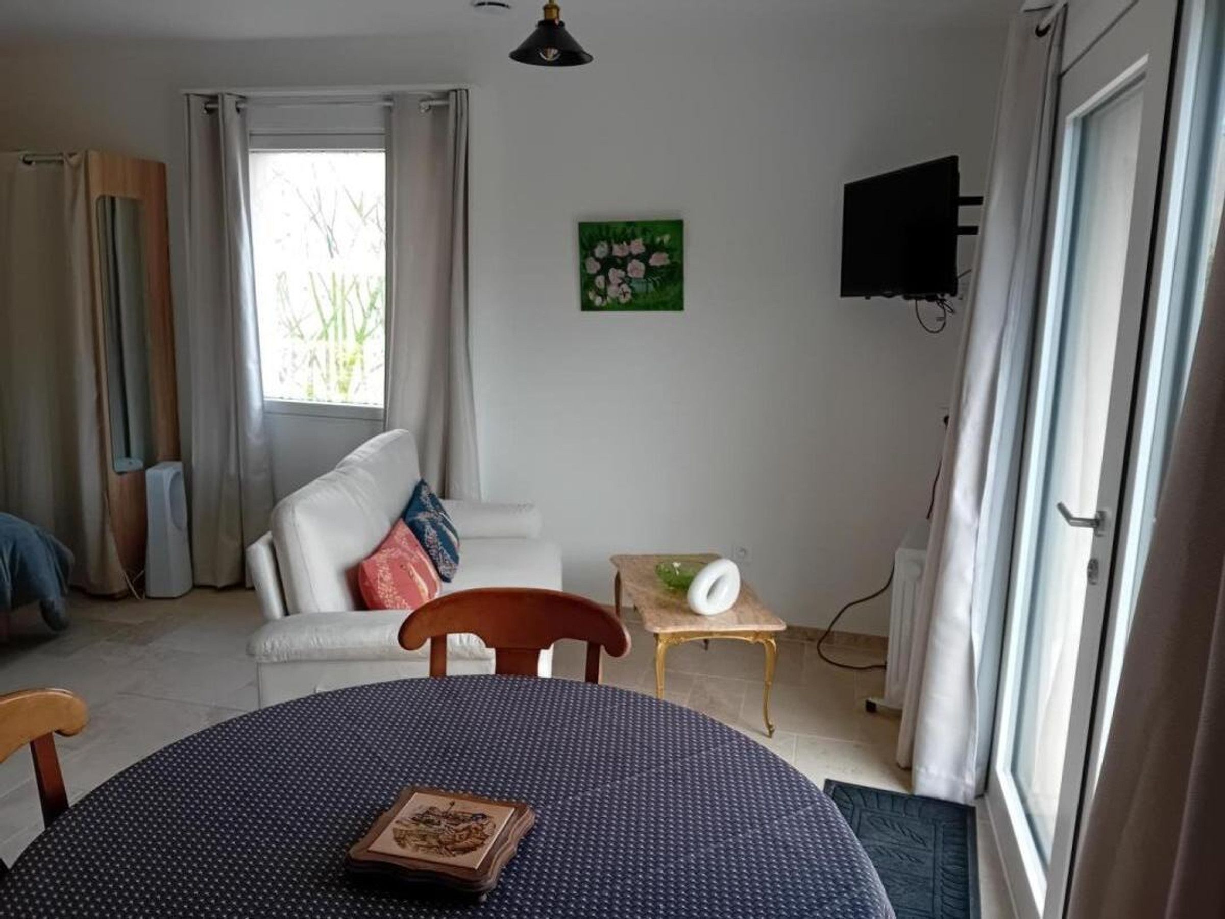 Ein Wohnzimmer mit einem weißen Sofa, einem runden Tisch mit gemustertem Tischläufer, einem an der Wand befestigten Fernseher und einem Fenster mit Vorhängen.
