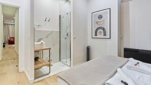 Une chambre à coucher moderne avec un lit, une table de chevet, une cabine de douche en verre et une œuvre d’art abstraite encadrée accrochée au mur.