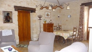 Un salon comprenant un canapé, un fauteuil et une table à manger avec des chaises. On y trouve une porte en bois, une horloge murale et un cadre photo accroché au mur.