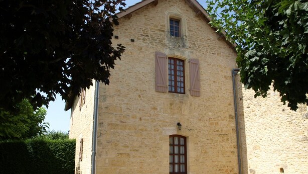 Un bâtiment en pierre doté d’une porte et de fenêtres, entouré de gravier et d’arbres.