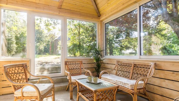 Eine überdachte Terrasse mit Korbmöbeln und großen Fenstern mit Blick auf einen Garten.