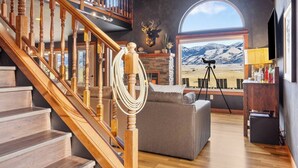 A spacious living room with a wooden staircase, a fireplace, a large window with a mountain view, and a leather couch. - Yellowstone Fun House | Hot Tub, Game Room + 360° Views (Emigrant)