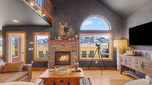 A spacious living room with a stone fireplace, leather furniture, a mounted deer head, and large windows offering mountain views. - Yellowstone Fun House | Hot Tub, Game Room + 360° Views (Emigrant)