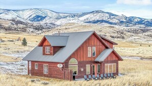 A wooden cabin with a sloped roof, a front porch with chairs, and a view of snow-covered mountains. - Yellowstone Fun House | Hot Tub, Game Room + 360° Views (Emigrant)