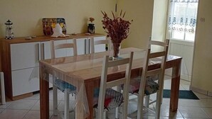 A dining area with a wooden table and chairs, a vase with dried plants, a painting on the wall, and a window with curtains. - Stone house near Utah Beach (Sainte-Marie-du-Mont)