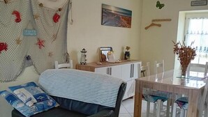 A living room with a black leather sofa, a dining table with chairs, a small kitchen area, and a decorative wall hanging. - Stone house near Utah Beach (Sainte-Marie-du-Mont)
