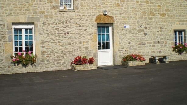 A stone building with a white door and windows, some with shutters. There are flower boxes with pink flowers on the ground level. - Stone house near Utah Beach (Sainte-Marie-du-Mont)