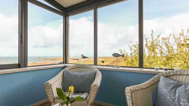 A conservatory with wicker furniture, a table with a teapot, and a view of the sea and beach. - Seaside House with Fireplace and Flowered Garden (La Hague)