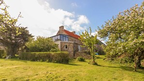 A stone house with a red roof, surrounded by a well-maintained lawn and mature trees. - Seaside House with Fireplace and Flowered Garden (La Hague)