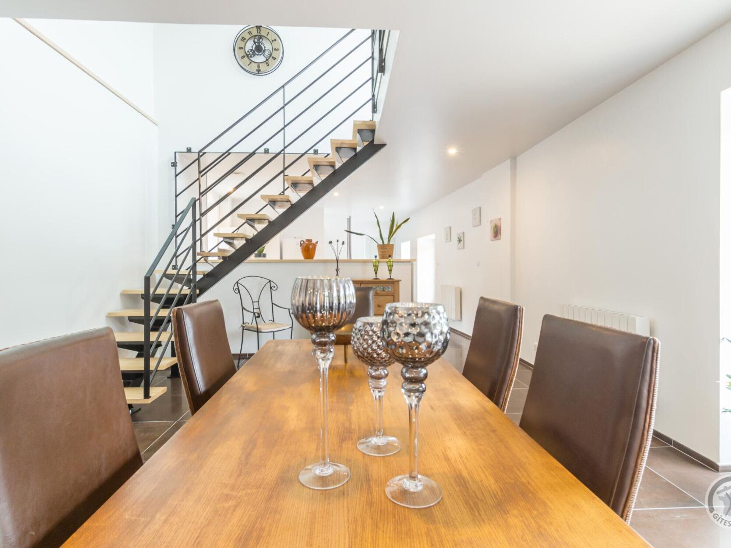 A dining area with a wooden table, six chairs, and three wine glasses. A staircase with a metal railing and wooden steps is visible in the background.