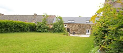 A stone building with a grassy yard, a hedge, and a large leafy plant in the foreground.