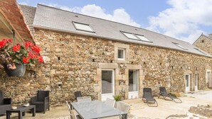 A stone building with a patio area featuring a table and chairs, a hanging flower pot, and a view of the sky. - Charming rental in La Hague with private garden, pets allowed (La Hague)