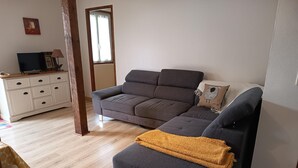 A living room with a grey sofa, a smaller grey ottoman, a wooden floor, a white cabinet with drawers, and a window with curtains.