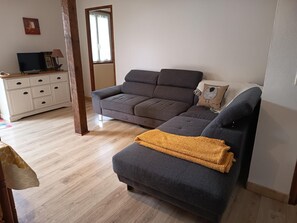 A living room with a grey sofa, a smaller grey ottoman, a wooden floor, a white cabinet with drawers, and a window with curtains.