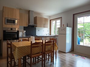A kitchen with wooden cabinets, a microwave, stove, and a refrigerator. There is a dining table with chairs and a window with a view of greenery.