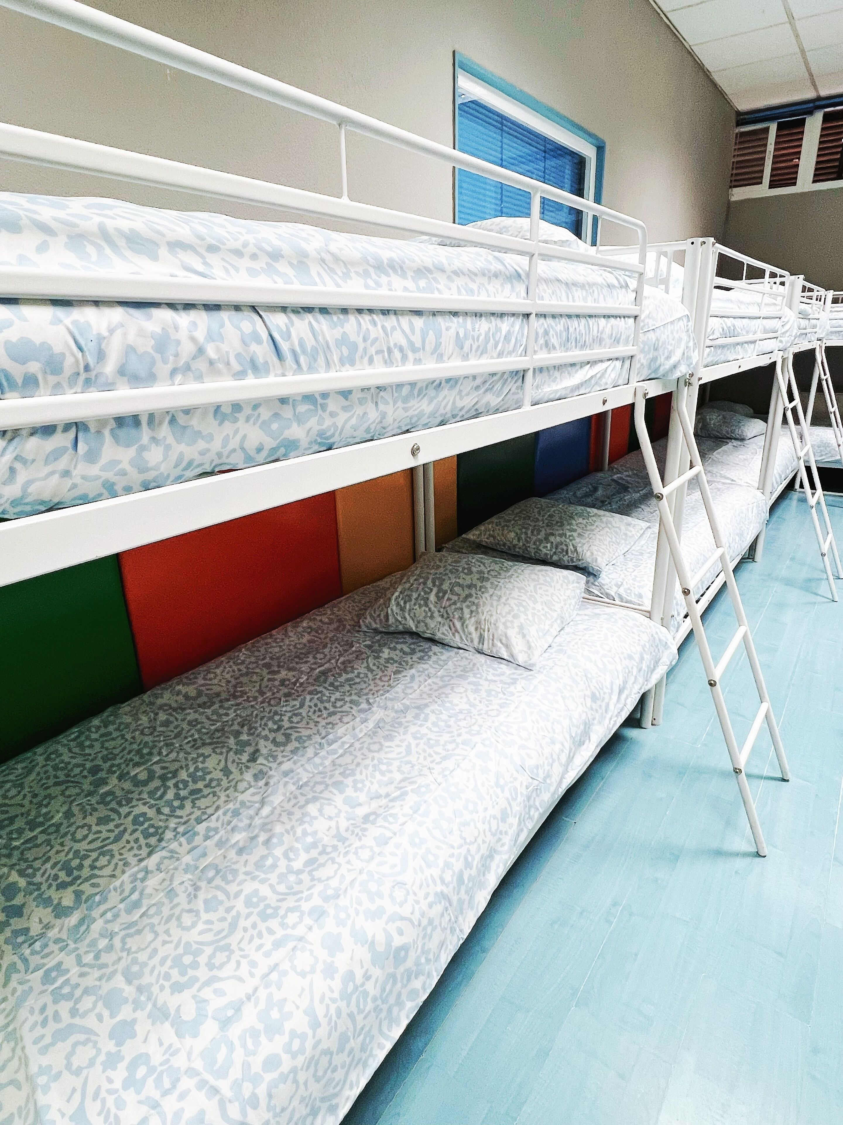Tweepersoonsbed met beddengoed met een patroon, een raam met zonwering en een deur met een blauwe omlijsting.