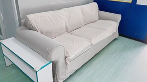 A light-colored sofa with a matching bench in a room with a blue accent wall and a window with blinds. - Hostel Las Rosas (Madrid)